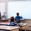A teacher stands to the left of a child and observes while the child writes on the chalkboard.