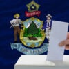 A hand deposits a ballot in a ballot box, with the Maine state flag in the background.
