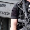 An armed U.S. Customs and Border Protection agent stands outside with a gun strapped to him.