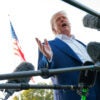 Donald Trump stands and holds his right hand out while he is surrounded by microphones.