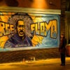 A man stands in front of a mural of George Floyd.