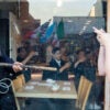 A police officer in riot gear hold a baton with two hands pointed at a protester who is saying something while making an obscene gesture with two hands.