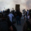 Shot from behind, a swarm of anti-ICE, anti-Trump protesters moves toward police in a street filled with smoke.