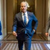 Senate Majority Leader John Thune strolls down a Senate corridor, flanked by two other people.