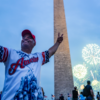 A man in an "America" jersey takes a selfie in front of the Washington Monument with fireworks and a crowd in the background.