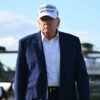 President Donald Trump in blue jacket, white shirt and white MAGA hat walks toward camera, with Marine One behind him.