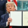 President Donald Trump, sitting at his Oval Office desk, holds up a signed Executive Order.
